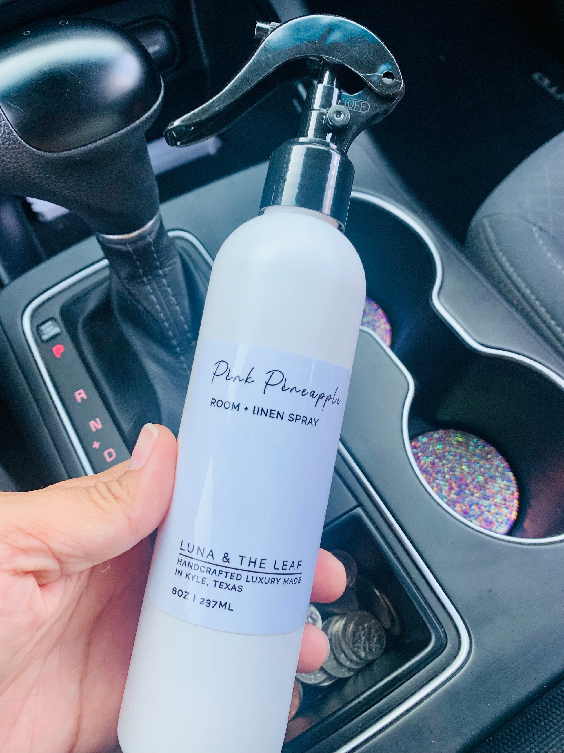 Hand holding a white bottle of Room Spray in the scent Pink Pineapple above the middle console in a car.