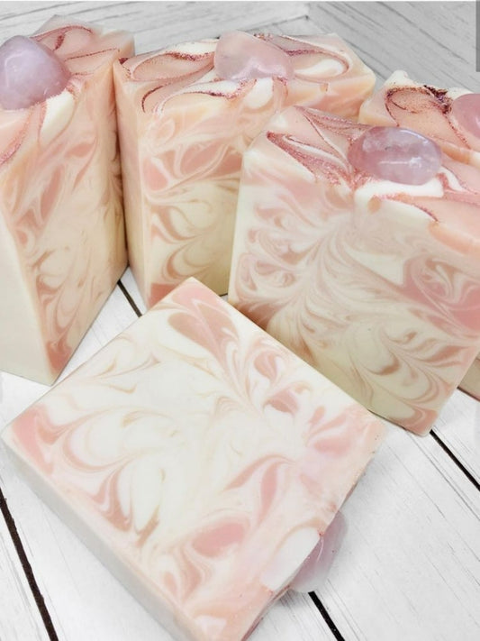 5 white and pink swirled soap bars topped with rose gold eco glitter and a rose quartz stone. One bar is lying down in front of the others. The soap bars are on a white wooden background. 