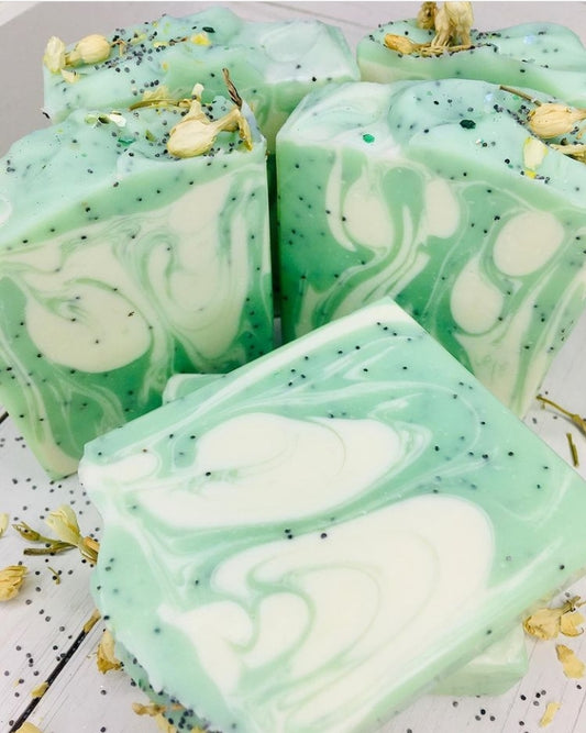 5 light green and white swirled soap bars with imbedded with poppy seeds. There are poppy seeds, jasmine flower buds, and eco glitter on the tops of the soap. The bar at the front is lying on its back. The soap bars are on a white wooden background with poppy seeds and jasmine flower buds scattered around the bars. 
