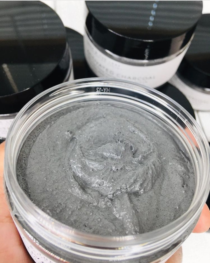 Clear containers filled with a grey creamy looking substance. The containers have black lids and a white label that says Activated Charcoal and tea tree facial sugar scrub. A hand is holding one of the containers without a lid. The grey contents of the container are close to the camera so you can see the texture of the scrub.