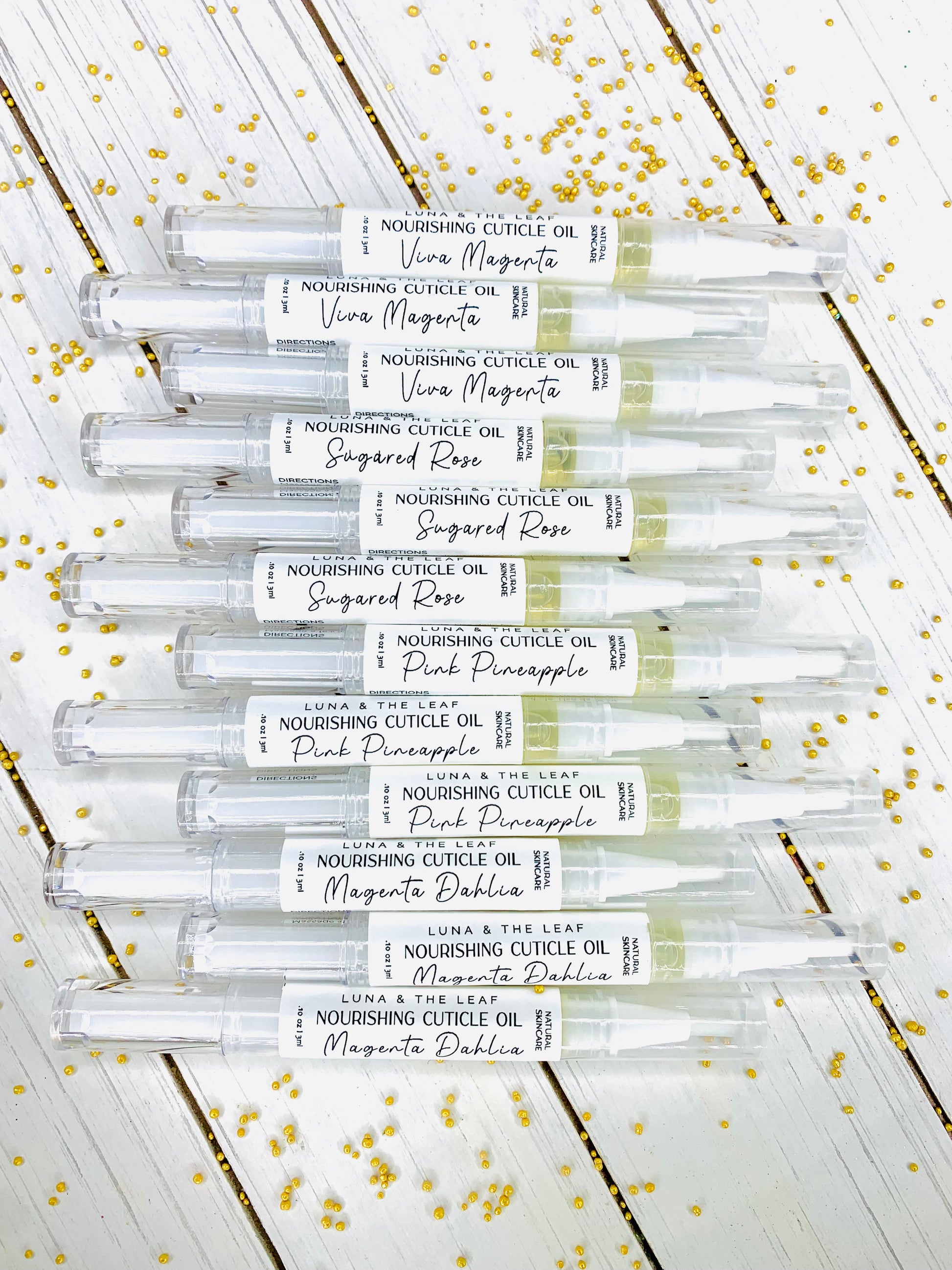 12 plastic filled cuticle oil pens laying horizontally on a white wood background. Small gold beads are scattered around the cuticle oil pens. The pens are labeled, the label reads Luna & The Leaf nourishing cuticle oil, natural skincare, .10 oz, 3 ml.
