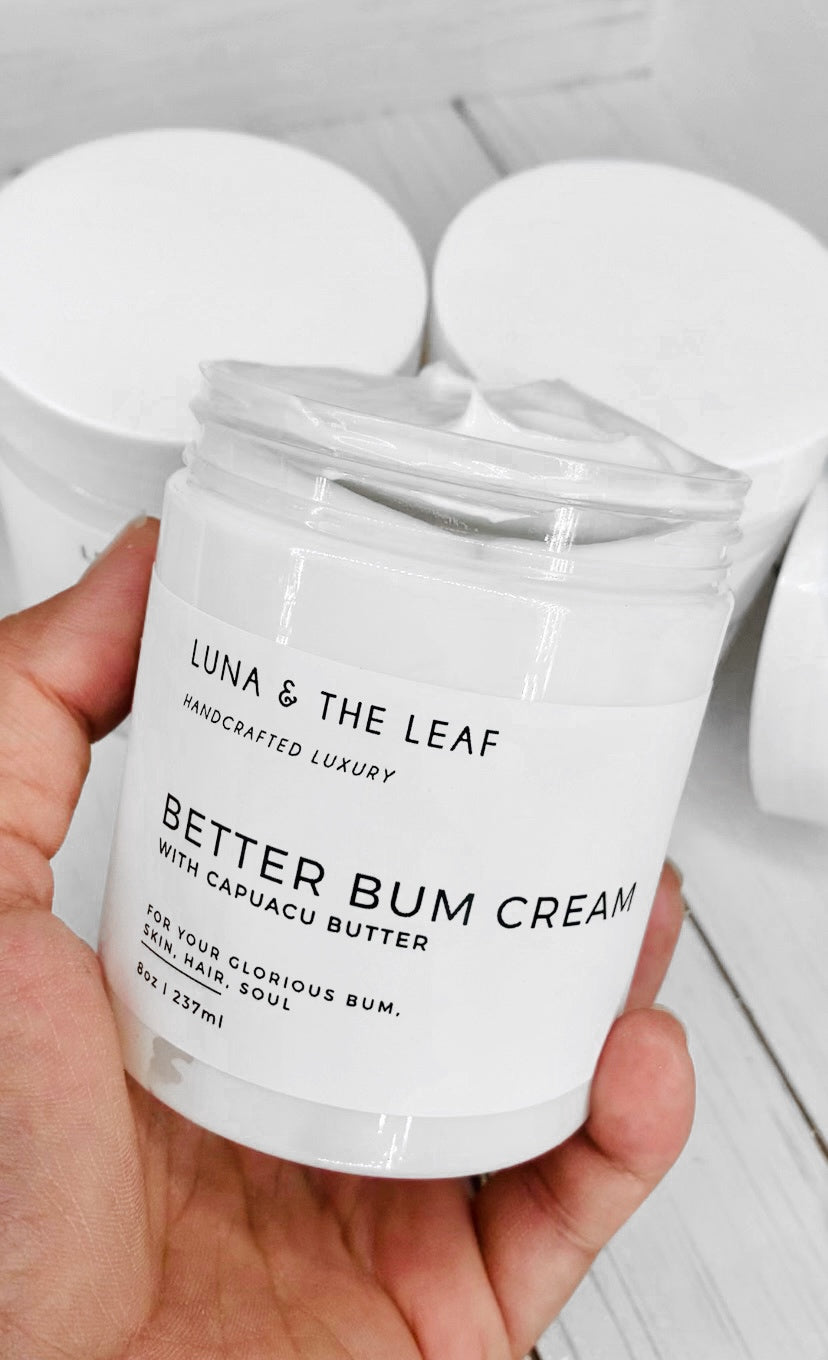 White cream in a container with a white label that reads Better Bum Cream with Cupuacu butter. It is being held by a hand with two more containers in the background. It is on a white wood background