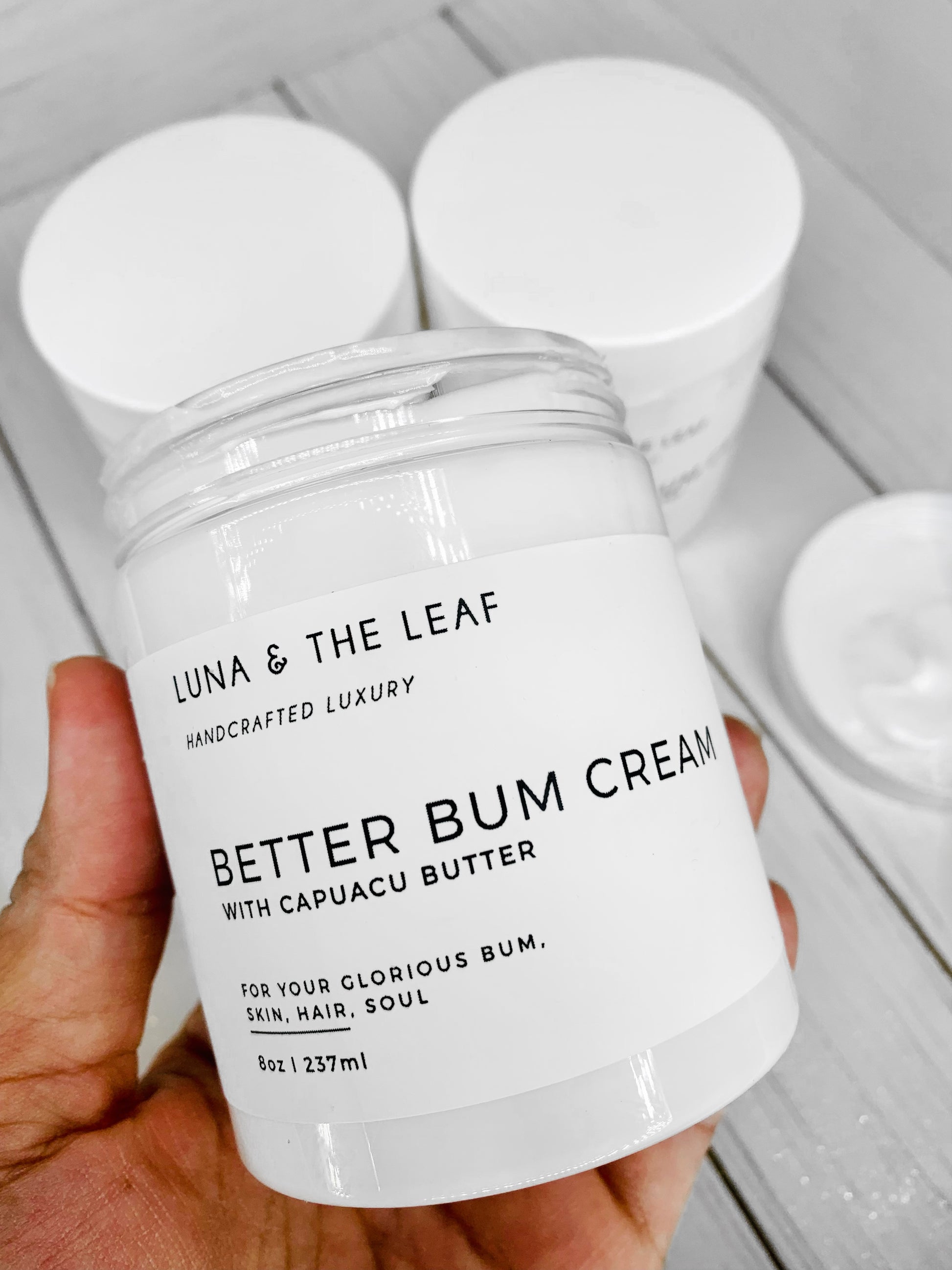 White cream in a container with a white label that reads Better Bum Cream with Cupuacu butter. It is being held by a hand with two more containers in the background. It is on a white wood background.