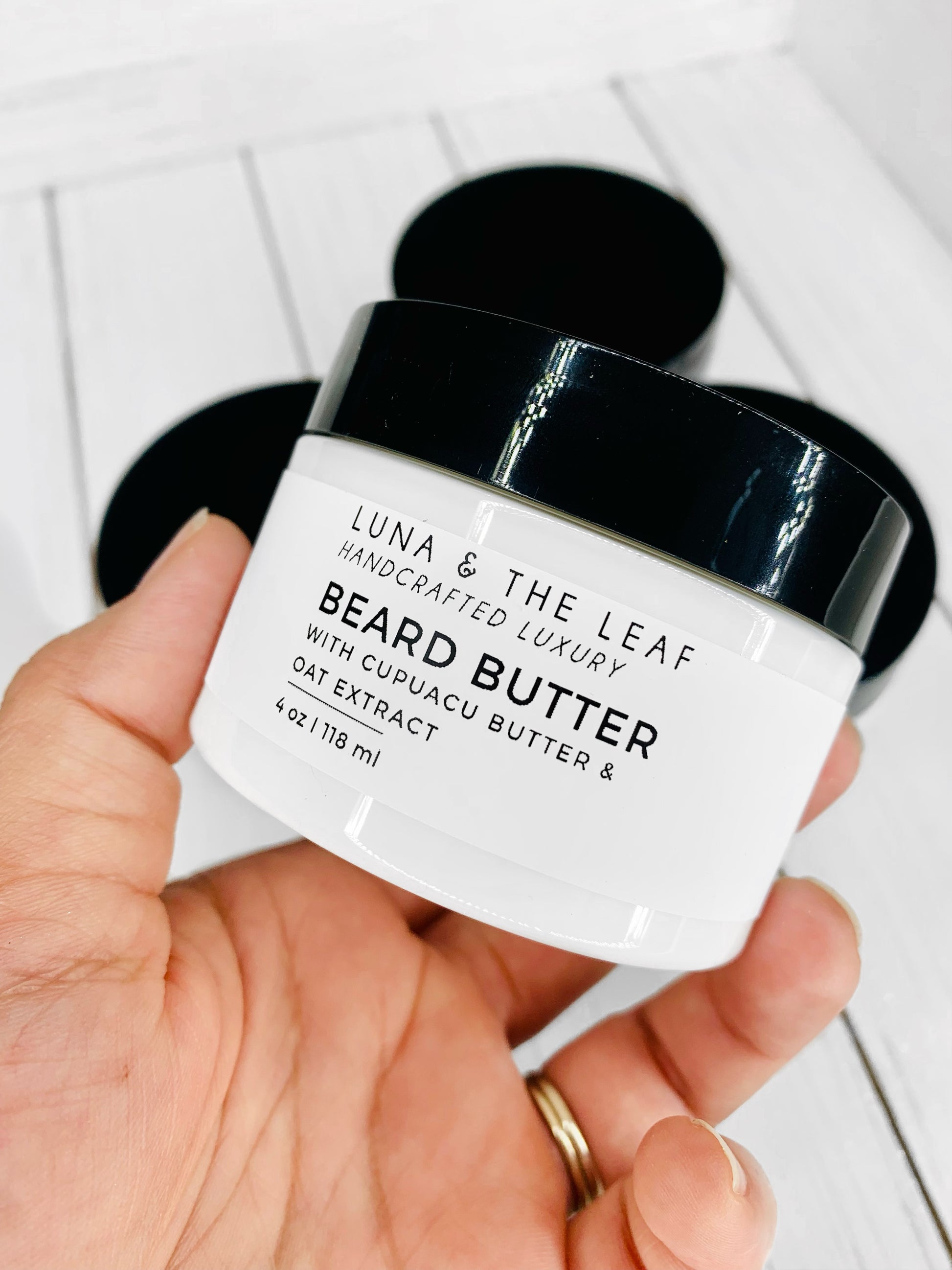 White cream in a container with a white label that reads Beard Butter. Container is being held in a hand. It is on a white wood background with three other containers in the background.