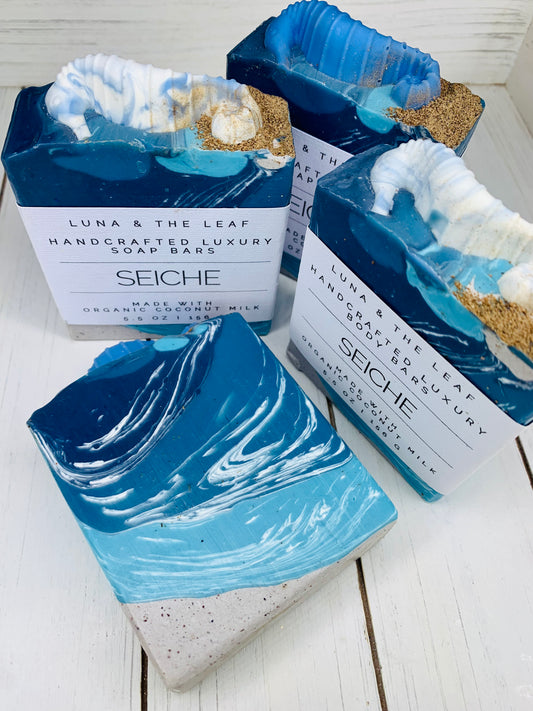 4 soap bars, one is lying at the front with no label to show the design. The other soap labels read Luna & The Leaf handcrafted luxury soap bars, seiche, made with organic coconut milk, 5.5 oz, 156 g. Each bar is colored with a gradient of white to dark blue and topped with a soap embed in the shape of a seahorse. The soap bars are on a white wooden background. 