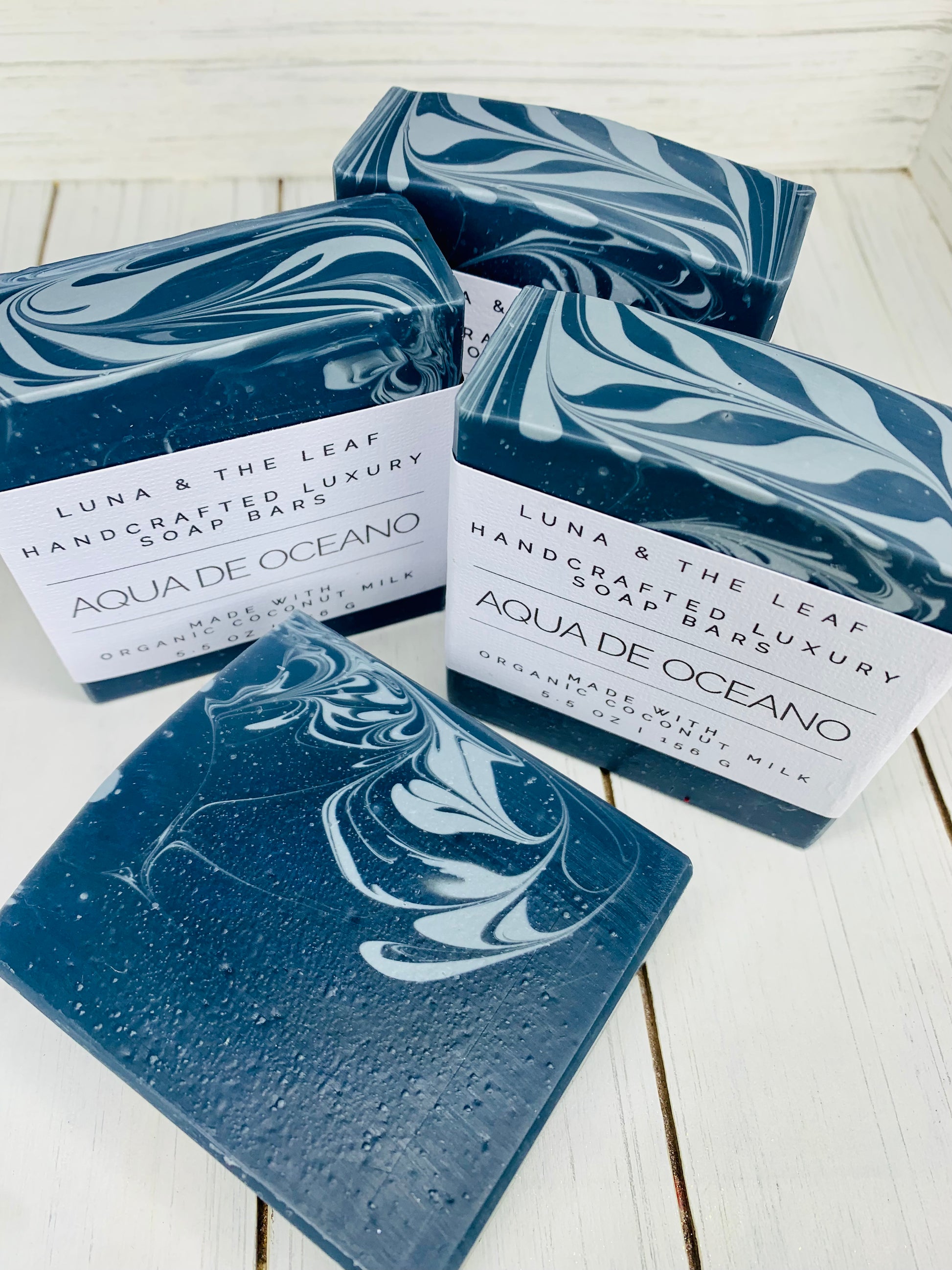 4 navy blue soap bars with a white swirl on one side and navy blue and white chevron pattern on top. Three are standing up with white cigar band labels that say Aqua De Oceano and one is laying down in front with no label. They are on a white wooden background.